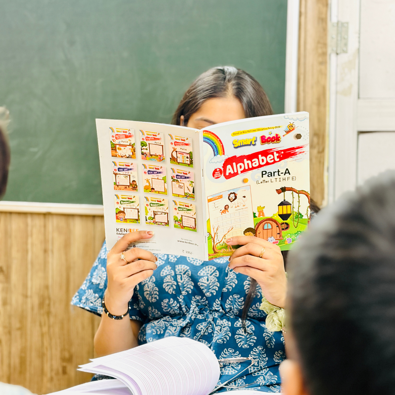Teacher holding Kenbee Book Alphabet Part-A educational workbook in a classroom