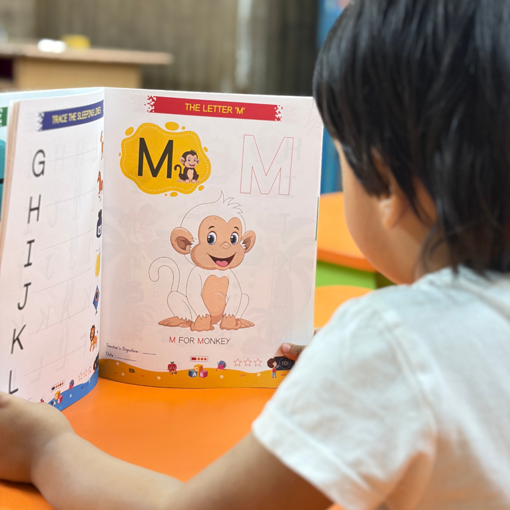 A young child looking at an open page of Kenbee Book | Patterns with Alphabet Part-A Beginner featuring a large cartoon monkey illustration for teaching the Letter M.