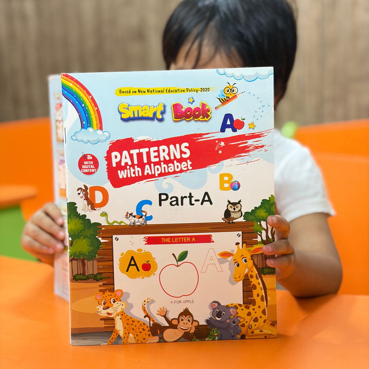 A young child holding up the front cover of Kenbee Book | Patterns with Alphabet Part-A Beginner, highlighting the vibrant cartoon animal illustrations and colorful "Smart Book" branding.