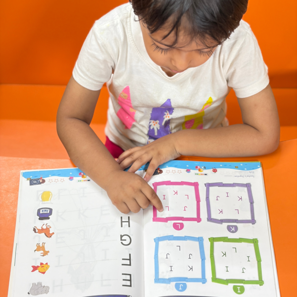 A child pointing at an interactive letter-finding puzzle page inside Kenbee Book | Patterns with Alphabet Part-A Beginner, featuring colorful box grids filled with mixed letters.