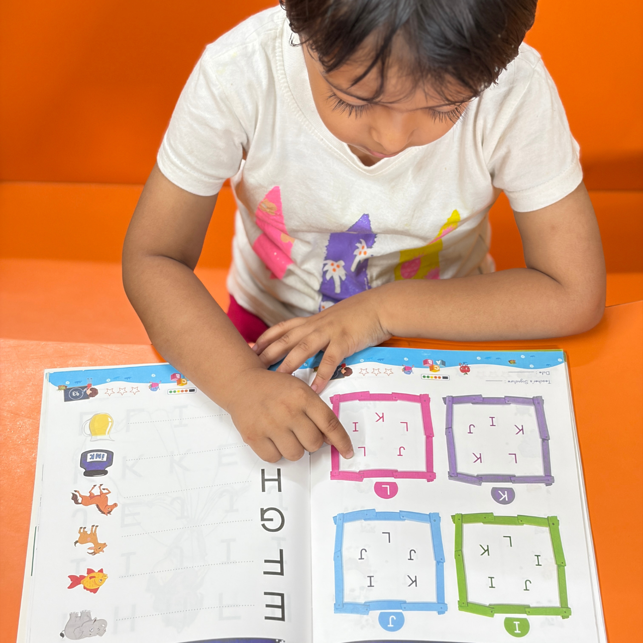 A child pointing at an interactive letter-finding puzzle page inside Kenbee Book | Patterns with Alphabet Part-A Beginner, featuring colorful box grids filled with mixed letters.