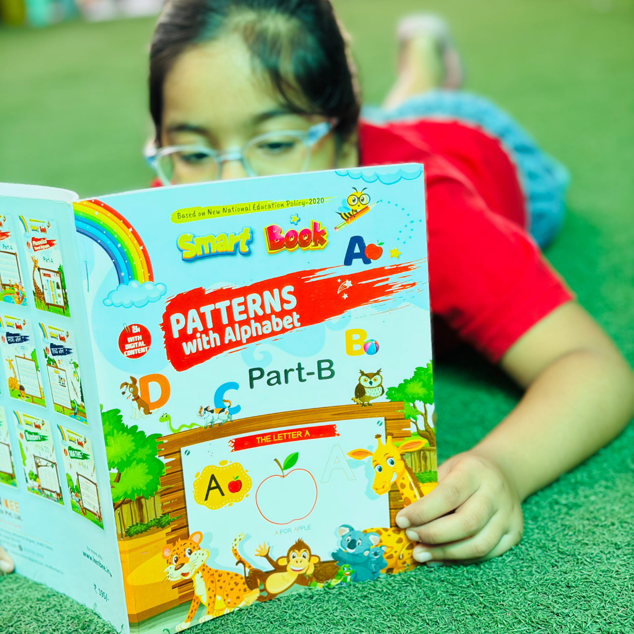 A young girl with glasses lying down and reading Patterns with Alphabet Part-B Beginner, focusing on the vibrant cartoon front cover featuring a giraffe, monkey, and leopard.