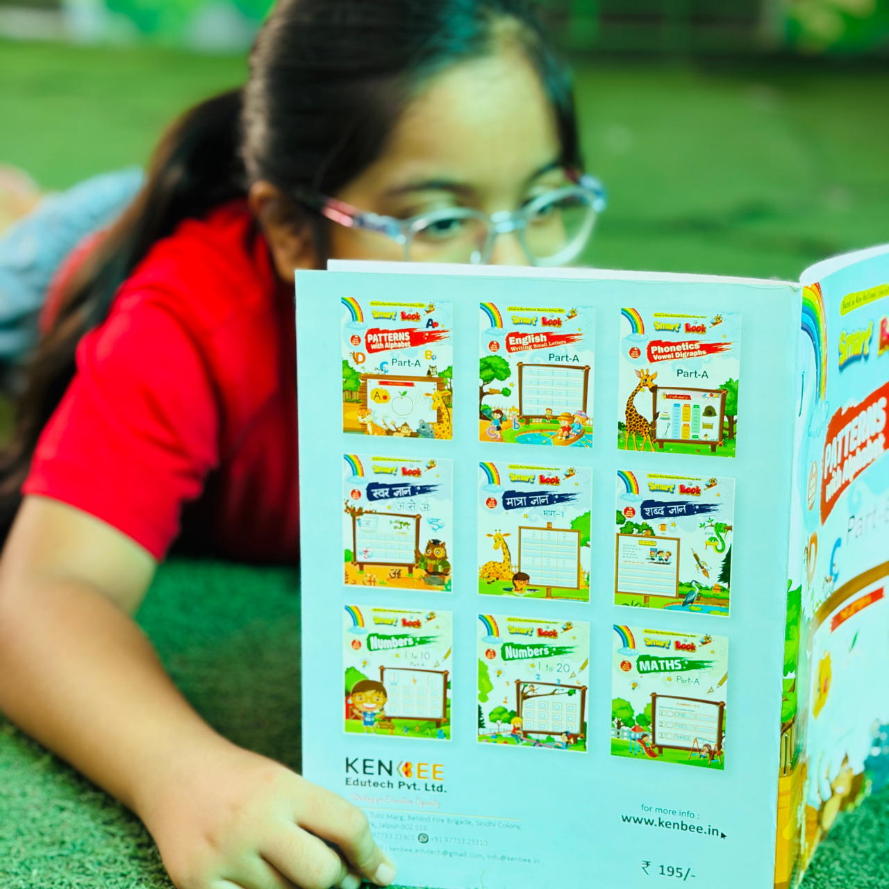A young girl lying down on a green surface while reading the back cover of Patterns with Alphabet Part-B Beginner, which displays an educational book grid catalog from Kenbee Edutech.