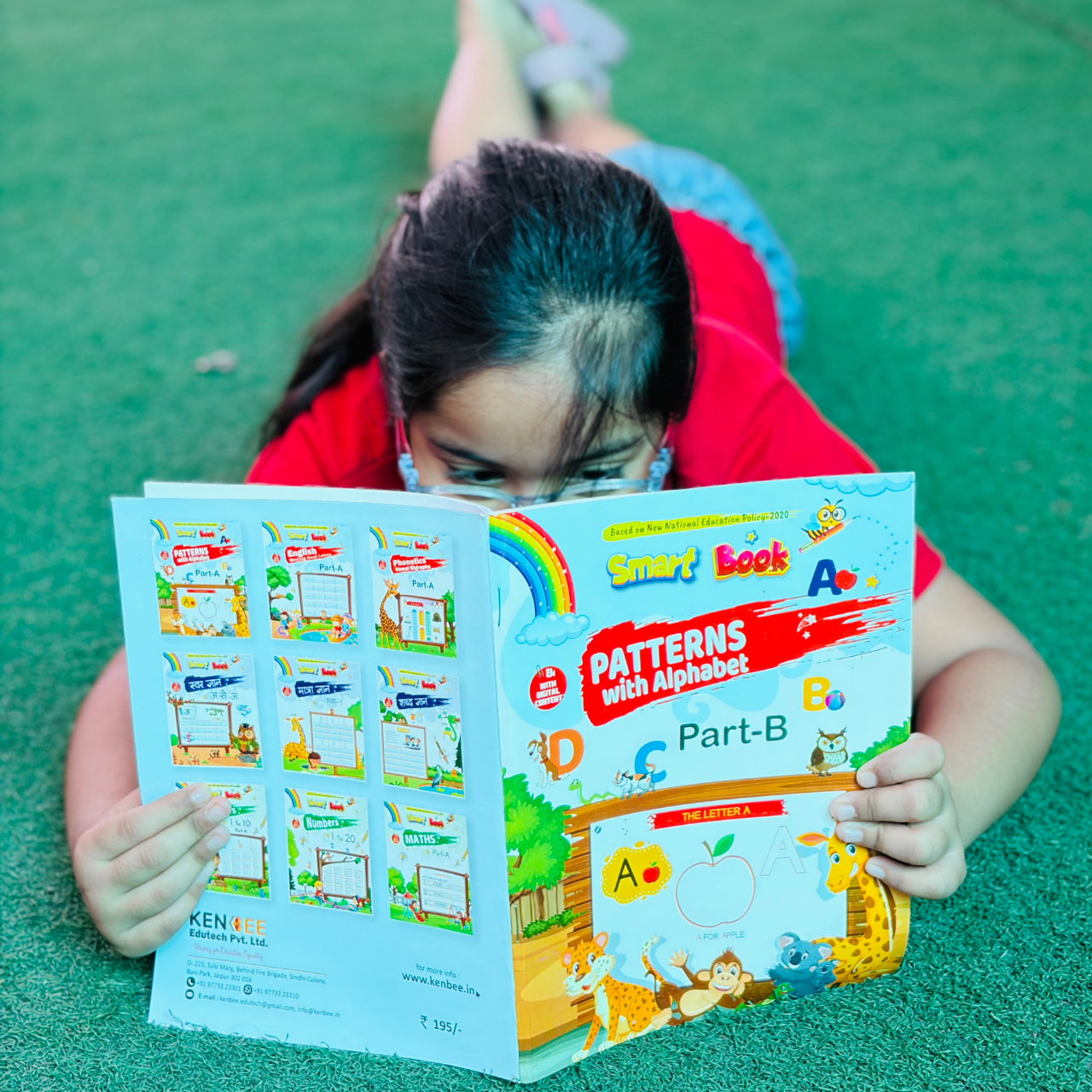 An overhead view of a child holding open Patterns with Alphabet Part-B Beginner, revealing both the colorful cartoon front cover and the product catalog on the back cover.